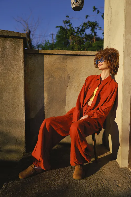 A young woman with curly hair sits against a concrete wall in a bold red suit, smiling softly as sunlight warms the urban courtyard ambiance