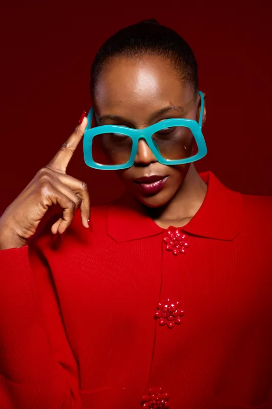 Stylish African woman with dark skin wearing large blue glasses and a red sweater touches her temple thoughtfully against a red background. Concept of fashion, confidence, and elegance.