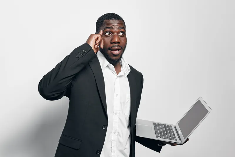 African American man with dark skin holding laptop and touching temple thinking about business or technology in formal suit on white background. Professional, focused and confident.