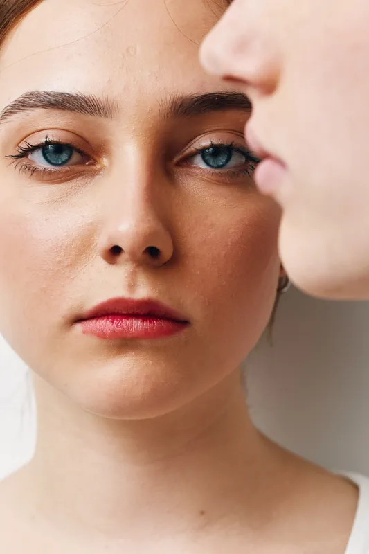 Portrait of young woman with blue eyes and red lips looking directly into camera with neutral expression, closeup of face and skin texture in natural light with blurred person nearby.