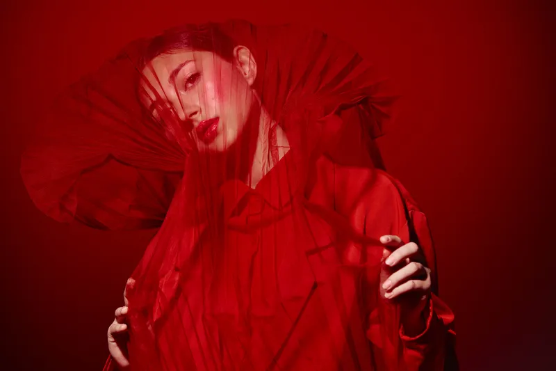 Fashion model with a dramatic red veil and matching outfit poses elegantly in a red studio. Creative beauty shot captures luxury, intense makeup, and sophisticated style.