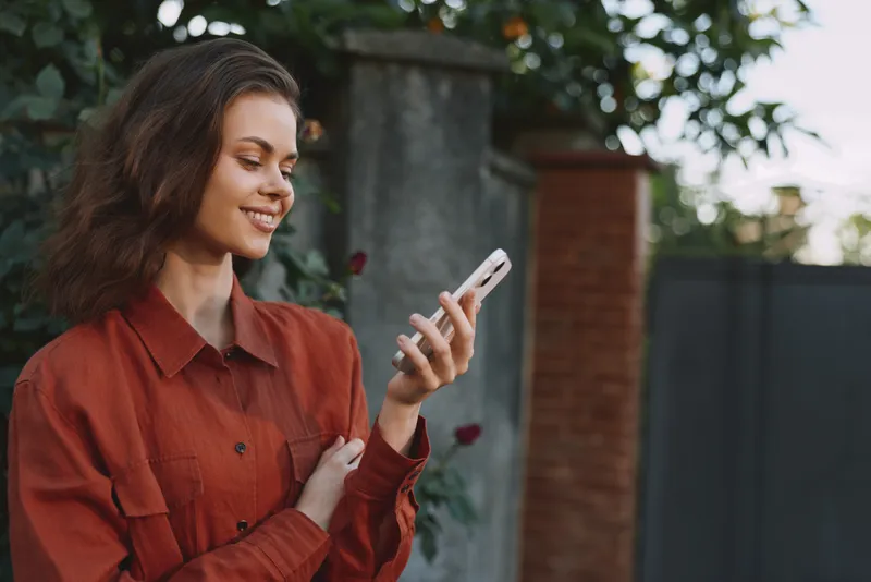 A smiling woman in a red shirt stands outdoors, looking at her smartphone with relaxed confidence as garden greenery frames the scene