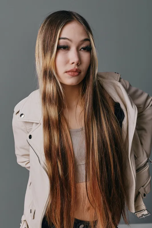 Young woman with long straight hair posing against gray background, wearing light jacket and neutral top. Fashion portrait of confident female model with natural makeup and relaxed expression.