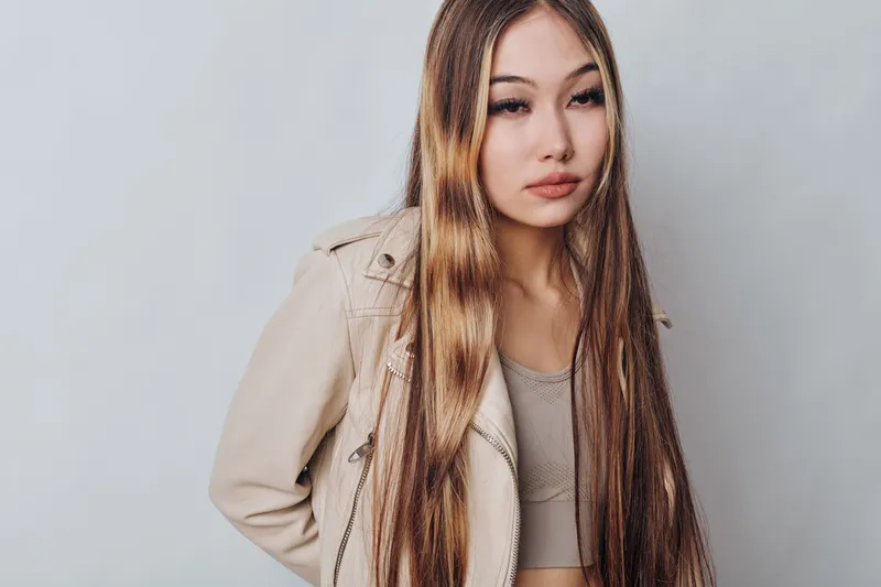 Young woman with long straight hair wearing beige jacket and top posing indoors against gray background with serious facial expression and natural makeup.