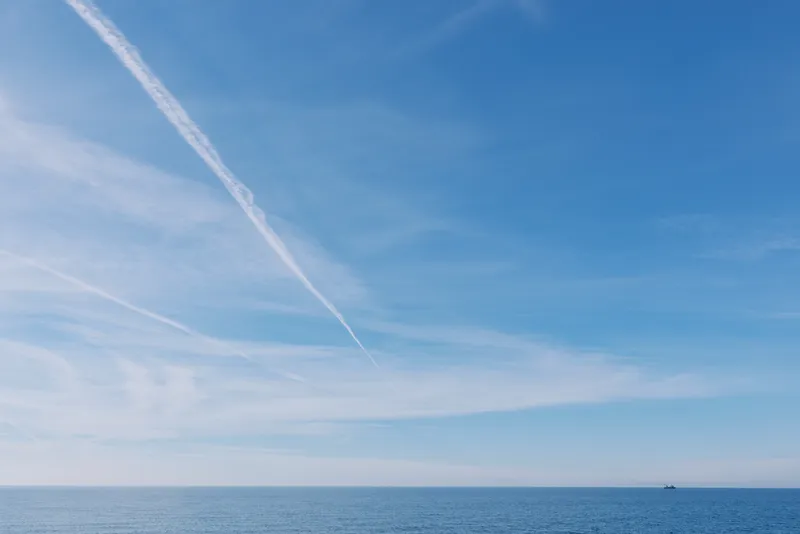 Sea and sky meet at a calm horizon with a bold contrail slicing across the blue expanse, creating a tranquil, expansive seascape under clear weather.