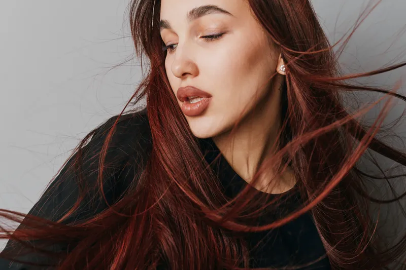Woman with long red hair and closed eyes posing in studio with soft lighting. Portrait of female model with natural makeup and flowing hair on neutral background.