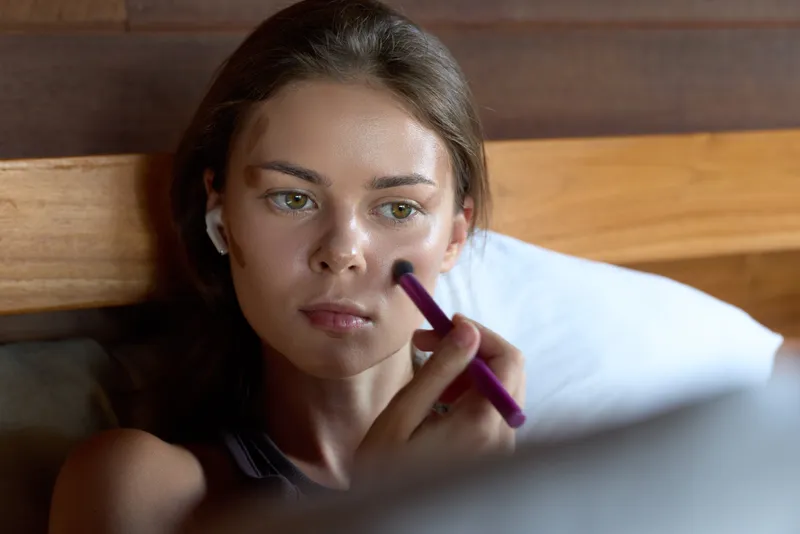 Makeup woman beauty morning lifestyle casual portrait applying cosmetics with focused expression, indoors under soft light, relaxed mood, closeup of face and makeupbrush in a cozy bedroom