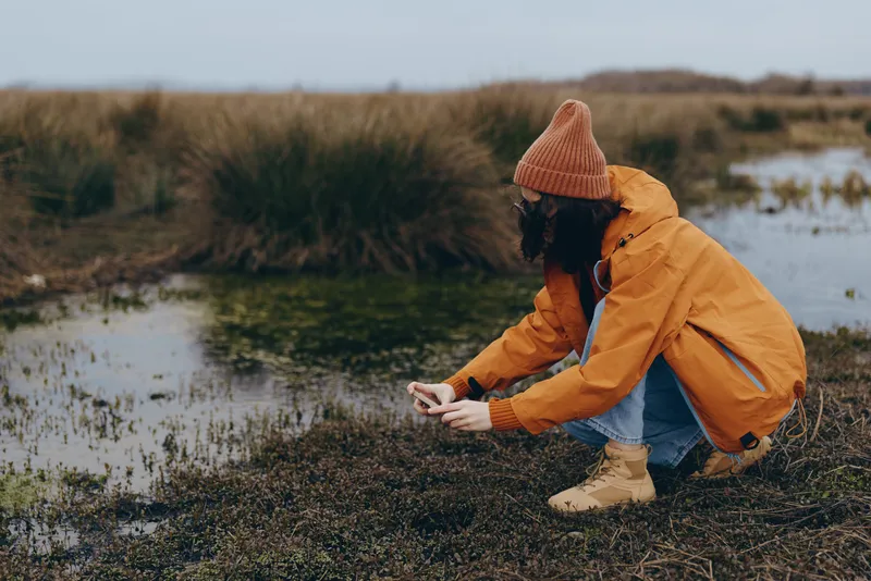 Woman outdoors in autumn lifestyle, wearing orange jacket and beanie, crouches near wetlands water edge, hands in soil, looking down, searching small plants, calm nature moment, daylight