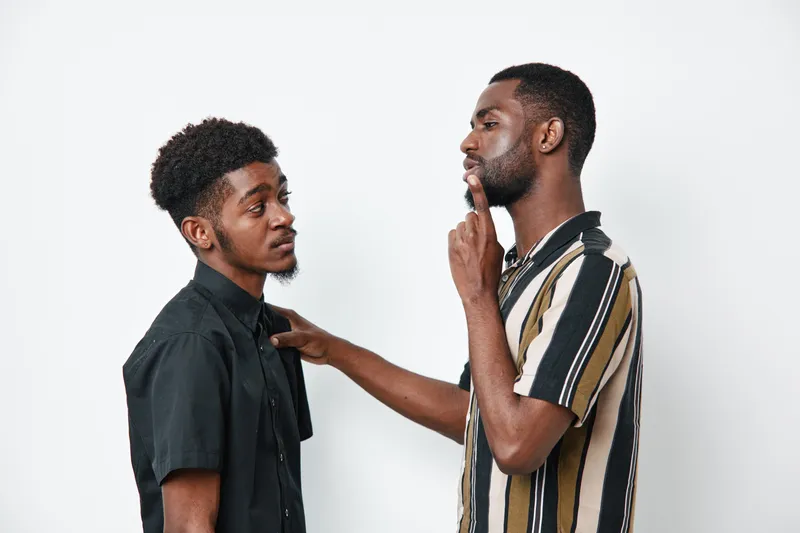 Two African American men with dark skin having a serious conversation in casual clothing against white background. One man is touching the other on the shoulder thoughtfully.