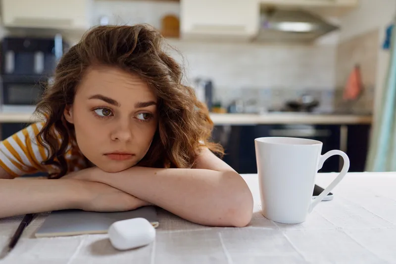 A woman sits at a kitchen table, resting her head on her arms, while a coffee mug sits nearby. The scene expresses sadness, fatigue, contemplation, loneliness, and a quiet home lifestyle.