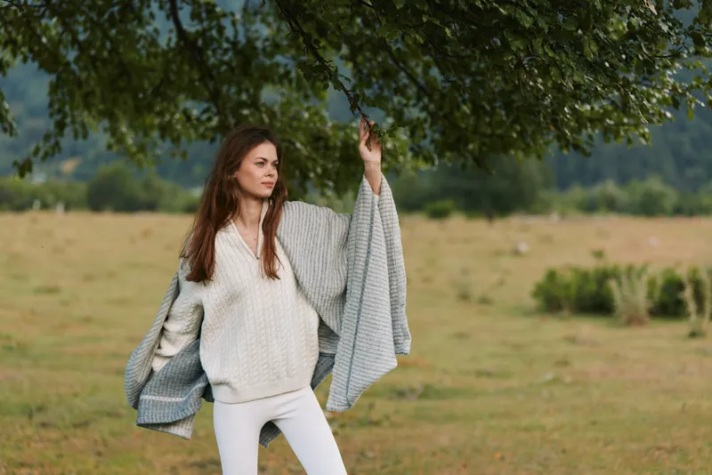 Woman standing in an open field under a leafy tree wearing a light sweater and white pants. Calm mood, casual fashion, nature backdrop, soft daylight.