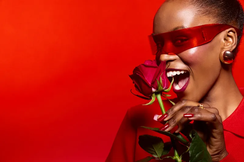 Smiling dark skinned African woman with red lips and nails holds red rose near mouth on red background. Concept of beauty, fashion, glamour, and elegance with vibrant color palette.