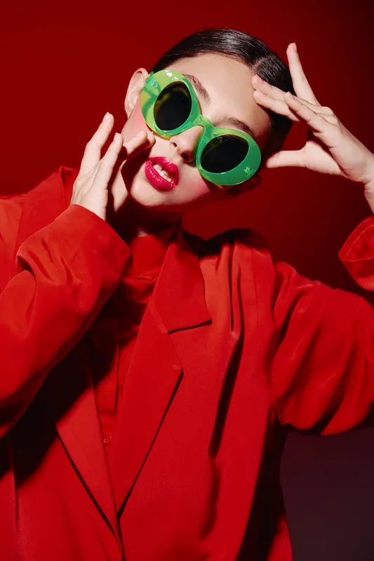 Woman in oversized green sunglasses and bright red blazer poses against a red background with glossy red lips. Bold beauty and fashion creative styling in the studio. Trendy glossy look.