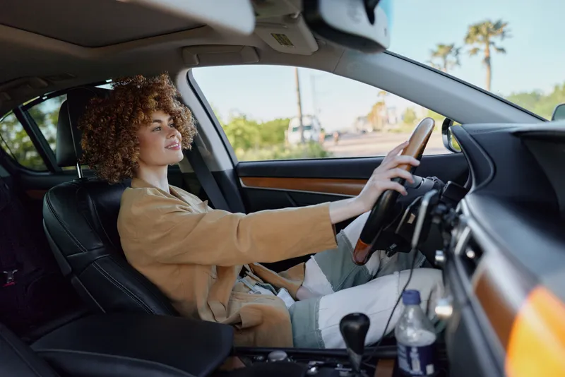 Cheerful woman driving a car, sunlit interior, relaxed posture, soft breeze through window, warm retro film color, natural tones, candid moment and calm outdoor light, distant city