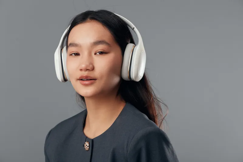 Young woman with long hair wearing wireless headphones standing against gray background with slight smile, listening to music or audio content in casual outfit indoors.
