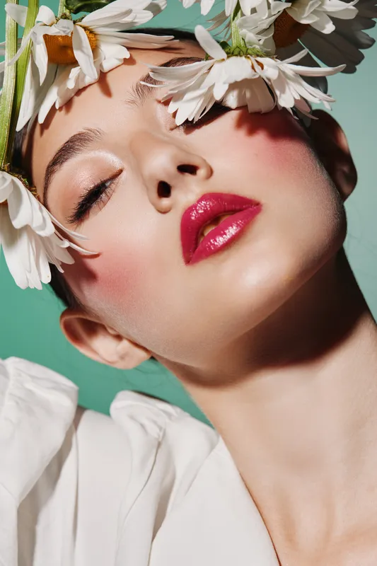 Close-up of woman with glossy red lips and vivid makeup surrounded by white flowers, eyes closed in a creative beauty fashion concept with soft lighting emphasizing skin texture.