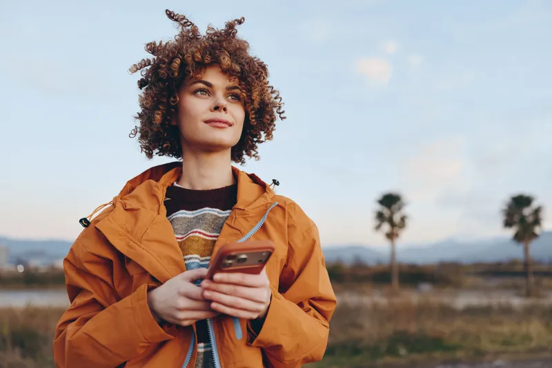 Cheerful woman outdoors with curly hair holding smartphone, orange jacket, looking at camera in sunlit field, relaxed lifestyle, soft breeze, natural colors, palms and distant sky