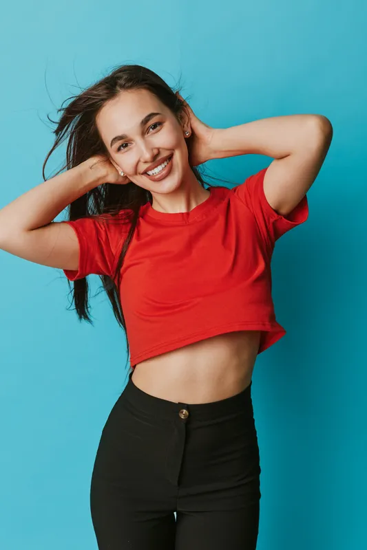 Young woman smiling and posing with hands behind head wearing red crop top and black pants against blue background. Casual style with confident and happy expression in studio.