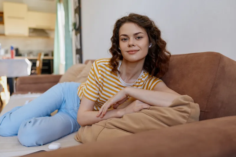 Young woman lounges on a beige sofa at home, wearing a striped yellow white tee and light blue jeans, with a calm smile and relaxed mood in a cozy living room.