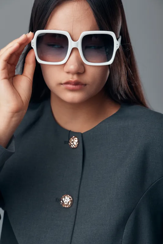 Woman wearing large white sunglasses and a dark blazer with decorative buttons, posing with eyes closed against neutral background in fashion portrait style.