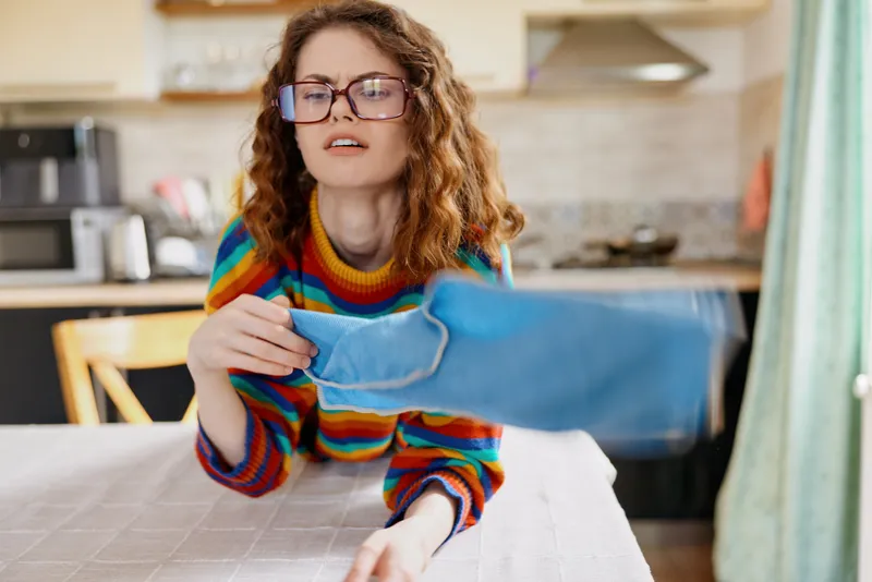 Smiling woman with curly hair wearing a colorful striped sweater irons a blue garment at a table in a bright kitchen, capturing a home lifestyle moment of daily chores and comfort today.