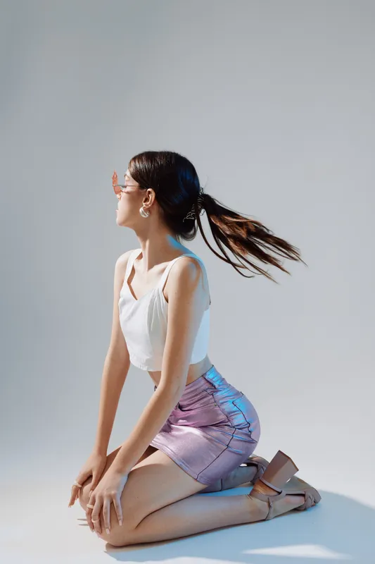 Young woman in fashionable clothes kneels on the floor looking to the side. She wears a white top and shiny purple skirt. Concept for woman beauty fashion and modern style.