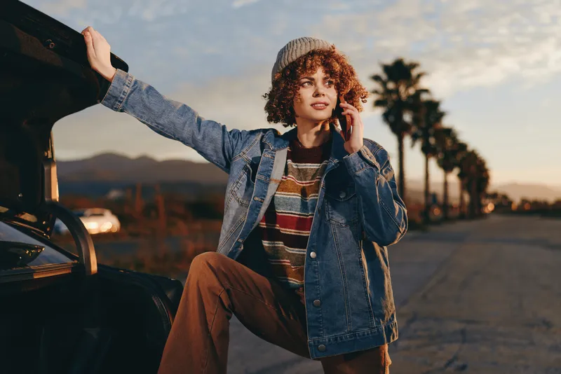 Woman lifestyle, denim jacket, curly hair, sitting on curb beside car, holding phone to ear, warm sunset light, casual street scene, relaxed travel mood outdoors