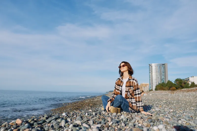 Woman sits on a pebble beach by the sea, wearing a plaid shirt and sunglasses, gazing toward the water with distant city buildings, a relaxed mood and natural light on the tranquil coast.