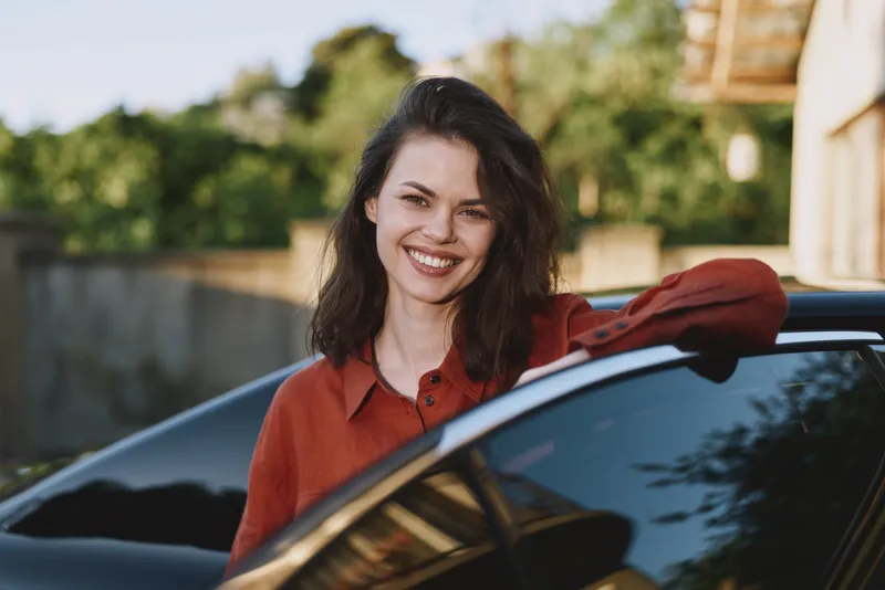 Confident woman leans on a sleek car outdoors, smiling warmly in a casual red shirt, capturing a relaxed lifestyle moment, modern neighborhood backdrop, bright natural light