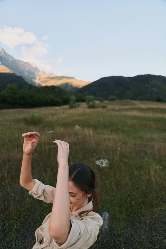 A young woman in a beige jacket raises her arms in a sunlit meadow with distant mountains behind, conveying a sense of freedom, calm and subtle joy as the breeze moves through tall grass and