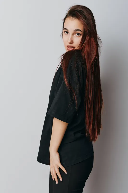Young woman with long brown hair wearing black t shirt and pants posing sideways on gray background. Female model with natural makeup and casual style looking at camera.