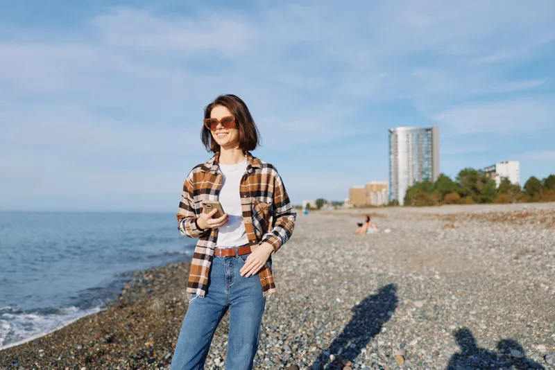 Casual woman on a pebble beach with phone, wearing plaid shirt, jeans and sunglasses, smiling under sunny sky with sea behind and urban buildings in distance, leisure moment outdoors.