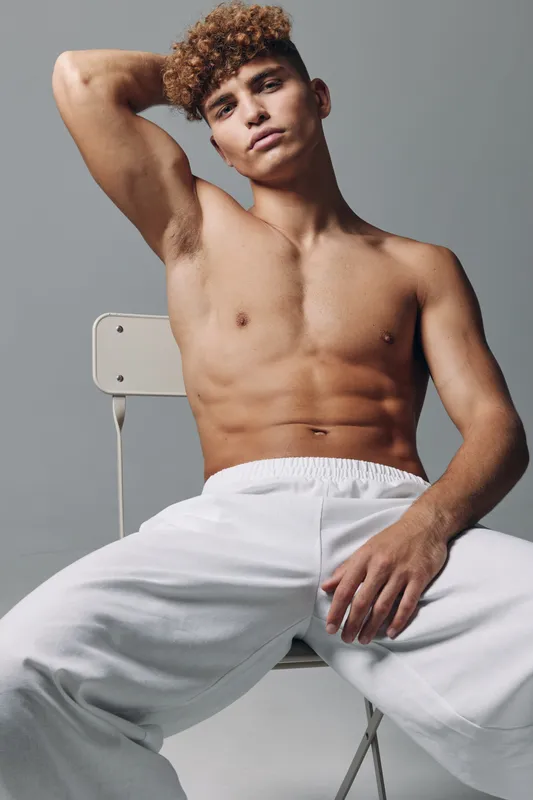 Young man with curly hair sitting on chair shirtless wearing white pants, posing with hand behind head and relaxed expression in studio with gray background.