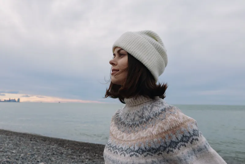 Woman in knitwear and a white beanie stands at a windy beach, looking toward the calm sea under an overcast sky, conveying a contemplative mood for nature and fashion.