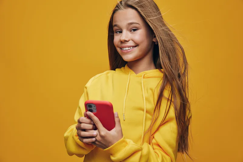 Teen girl smiling and holding smartphone with red case in yellow hoodie on yellow background. Happy young female using mobile phone for texting or social media communication.
