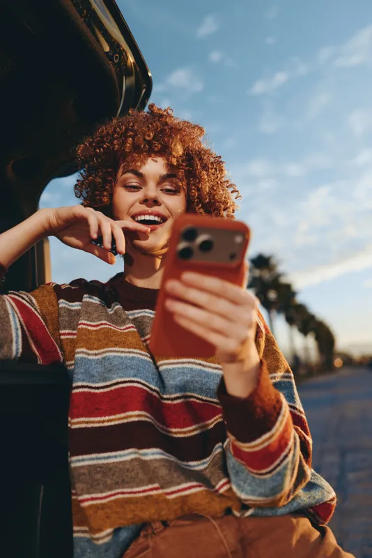Outdoor lifestyle woman with curly hair smiles while holding a smartphone for a selfie, striped sweater, casual fashion on a bright street portrait, joy and communication with friends