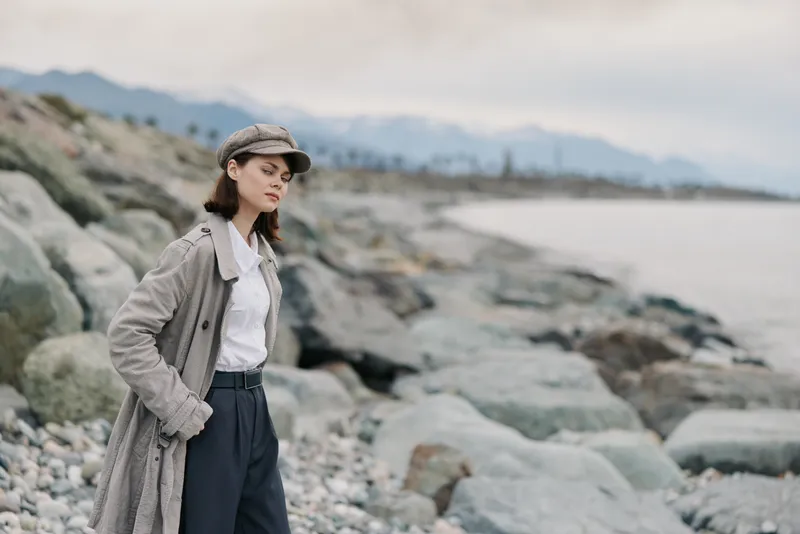 Young woman in a grey coat and hat stands on a rocky beach, looking thoughtfully into the distance. Overcast sky and calm sea create a serene coastal atmosphere.