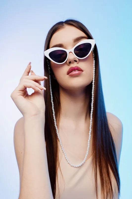 Young woman with long hair wearing white cat-eye sunglasses and pearl chain poses confidently against a light blue background. Concept of beauty fashion style.
