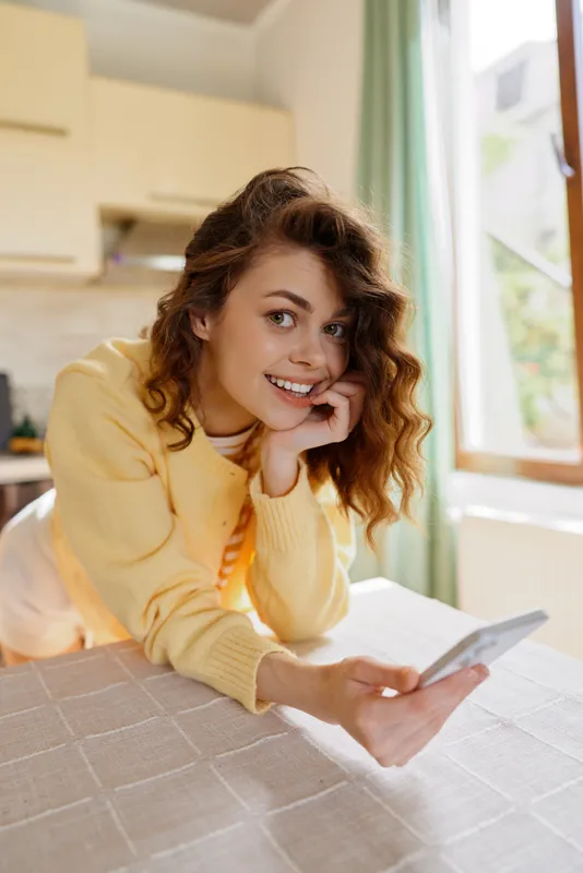 A young woman with curly hair relaxes at home, smiling softly as she leans on her hand and holds a phone for a casual lifestyle moment.
