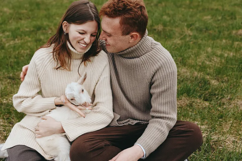 A young couple in cozy sweaters sits on grass outdoors while holding a small white baby goat, sharing a tender moment in a natural lifestyle setting during autumn.