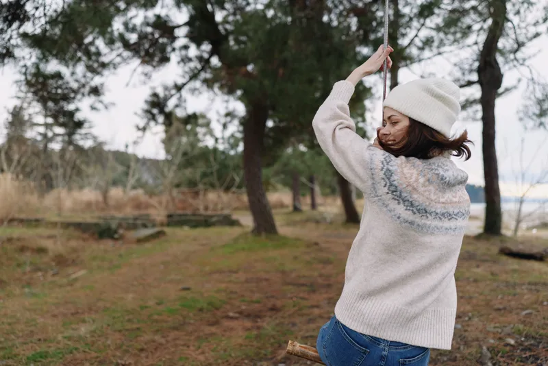 Woman in a cozy knit sweater and beanie stands outdoors in a park, arms raised, enjoying fresh air among trees and open space.