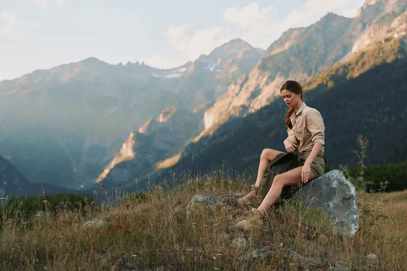 Young woman with long brown hair sits on a rock in a mountain meadow during golden hour, enjoying a peaceful moment in nature with a relaxed and thoughtful expression.