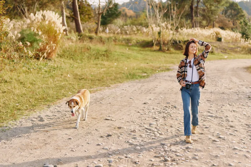 Woman walking her dog along a gravel path in a sunny countryside scene, casual attire, open landscape, and a peaceful outdoor moment of companionship and exercise.