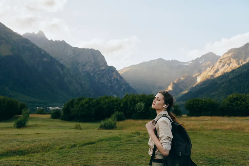 Young woman with a backpack stands in a mountain valley, gazing upward with a calm hopeful expression, amid green fields and distant peaks.