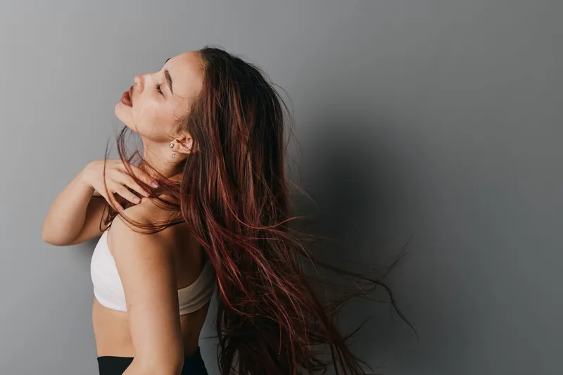 Woman with long hair in white top posing with closed eyes and hand near neck on gray background. Beauty portrait with natural makeup and flowing hair in studio lighting.