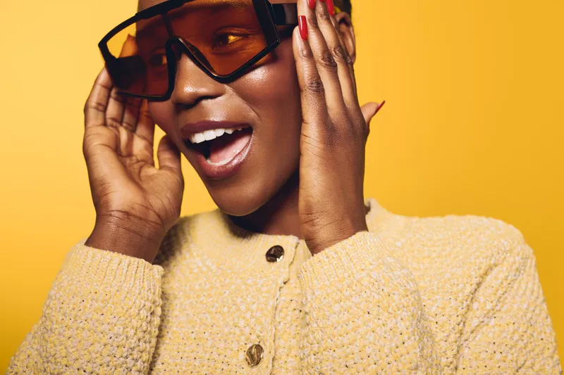 Happy African woman with dark skin wearing large sunglasses and beige knitted sweater smiles with open mouth on yellow background. Concept of fashion, style, and joyful mood.