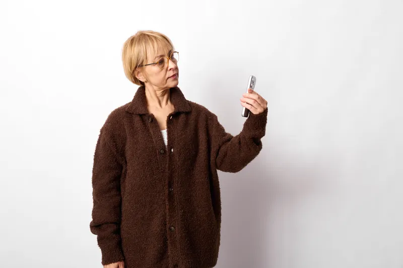Elderly woman portrait, wearing brown winter coat and glasses, holding smartphone for payment, looking at screen, neutral studio background, copy space, text