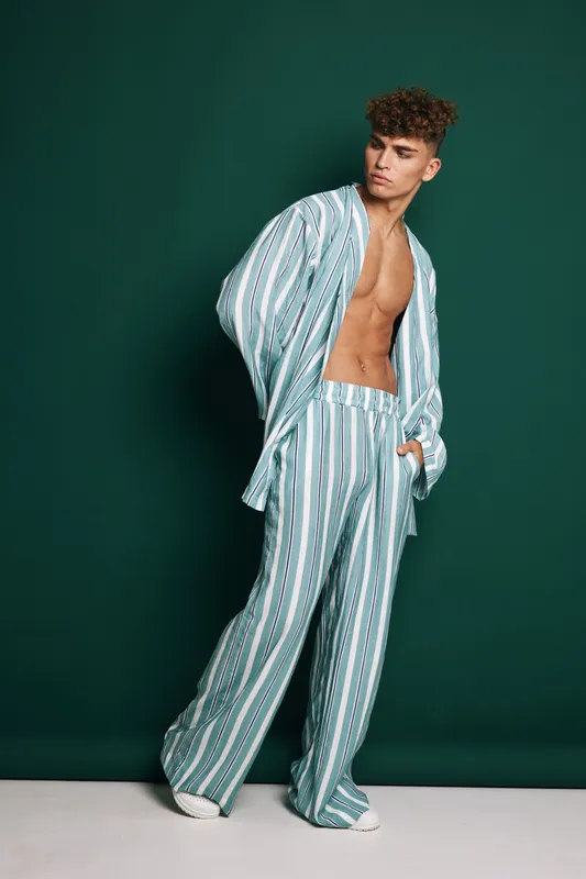 Young man with curly hair wearing striped loose pants and open shirt posing against green background. Fashion model with casual style and confident expression in studio setting.
