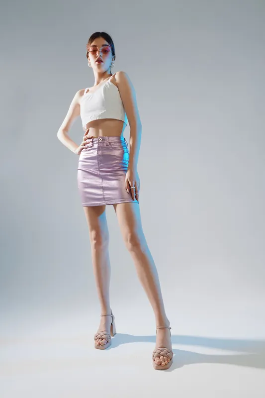 Young woman in a white crop top and shiny pink skirt poses confidently in high heels. Studio shot with soft lighting on a neutral background. Concept of woman beauty fashion.