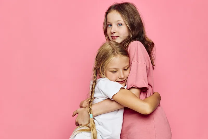 Two sisters hugging affectionately against a pink background. Older girl wearing pink holds younger girl in white shirt with braids.